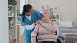 A caregiver in blue scrubs stands beside an elderly woman in a wheelchair, gently holding her shoulder.