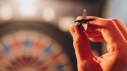 A hand ready to throw a dart with a blurred dartboard behind.