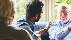 An elderly man with a cane speaks to a young man on a couch as an older woman observes in a Medbridge living room.