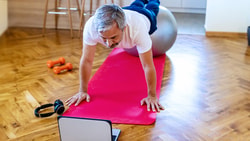 A person works out on a stability ball with a laptop, headphones, and dumbbells nearby on a red mat.