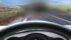 Blurry car windshield view of a road, grassy hill, and blue sky.