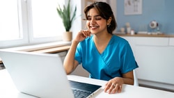 A person in blue medical scrubs smiles at a laptop, with a window and potted plant behind them.