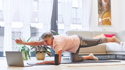 An older woman follows medbridge exercise videos on her laptop, balancing on a yoga mat.