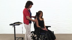 A woman helps adjust another woman's posture in a wheelchair near a table with electronic equipment, promoting Medbridge care.