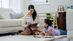 Occupational therapist sitting on the floor with a young girl, using a tablet during a therapy session at home.