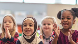 Children in a classroom smiling and clapping with blurred elements in the background.