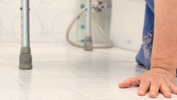 A person kneels on a bathroom floor with one hand down and a walker beside them.