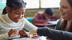 A student happily uses a tablet at a desk with their teacher in a Medbridge classroom.
