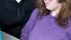 Two individuals smile at a blue tablet on the table, one wearing a black jacket and the other in a purple shirt, while using medbridge.