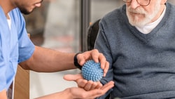 A Medbridge clinician in blue scrubs presents a textured object to an elderly man in a sweater.