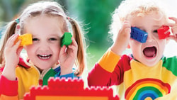 Two children in rainbow shirts hold colorful blocks to their eyes, smiling.