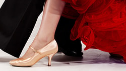 Feet of two dancers in motion, one in black shoes and pants, the other in gold heels with a red dress.