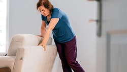 A woman in a blue shirt and dark pants holds a beige chair for support in a room, seen through a doorway.