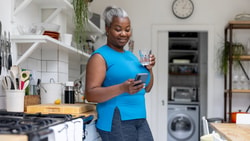 A woman in a blue top stands in a kitchen, holding a glass of water and looking at her phone, with kitchen appliances and plants behind her.
