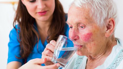 A Medbridge caregiver in a blue uniform helps an elderly person drink water.