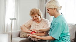 A nurse helps an elderly woman with hand exercises using a red ball on a couch.
