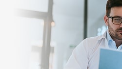 A person with glasses is thoughtfully examining a paper indoors.