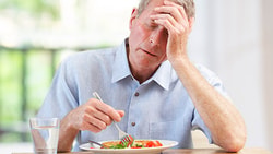 An elderly man in a light blue shirt sits at a table looking tired or stressed with food and water nearby.