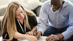 Clinician assisting an older adult with cognitive communication disorder during a writing exercise at home.