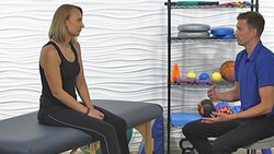 A woman sits in a therapy setting with exercise equipment behind her and a gesturing man from medbridge.