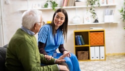 A nurse sits at eye level with an older patient, leaning in and listening during a calm bedside conversation.