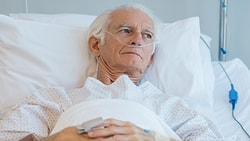 Elderly man in a hospital bed with an oxygen tube, wearing a gown, holding a device.