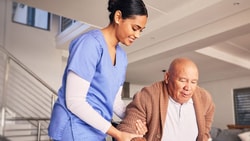 A Medbridge caregiver in blue scrubs helps an elderly man in a cozy living room.