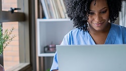 Home health clinician in scrubs reviewing OASIS documentation on a laptop, representing OASIS accuracy and training.