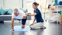 A physical therapist guides an older man through a bird-dog exercise for Medbridge training.