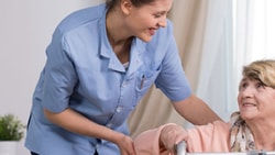 A Medbridge caregiver in blue helps a smiling elderly woman with a walker in a bright room.