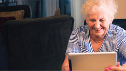 Smiling elderly woman with curly hair uses a tablet at a table with yellow flowers.