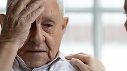 A distressed elderly man sits with his hand on his forehead, comforted by someone next to him touching his shoulder.