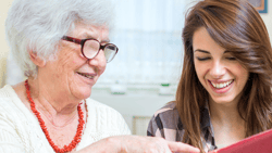 An elderly woman with glasses and a young woman with brown hair smile while reading a red book together.
