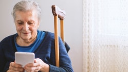 An elderly woman in a blue outfit focuses on her smartphone near lace-curtained window.