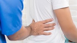 Someone in a blue shirt touches another person's back, against a white brick wall.