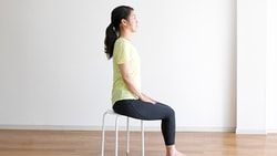 Someone in yellow and black sits on a stool in a minimalist room with a sofa, facing forward.