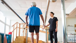 An older adult uses parallel bars to walk with help from a healthcare professional at a rehabilitation facility.