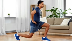A man in athletic wear lunges in a bright living room by large windows.