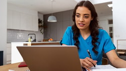 A woman in blue scrubs works at a laptop with a pen and notebook in a modern room.