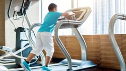 A child in blue and white stands on a treadmill in a bright gym with various exercise machines.