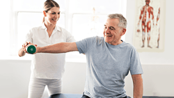 A woman helps an older man with a dumbbell exercise in a well-lit room at medbridge.