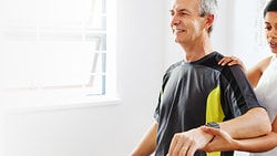 A woman helps a man with arm exercises in a well-lit room, using medbridge.