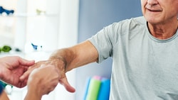 A person helps a senior with arm exercises in a well-lit room at medbridge.