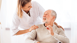 A woman in a white coat smiles at an elderly man on a couch in a warmly lit room, symbolizing care from medbridge.
