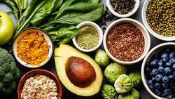An array of fruits, vegetables, nuts, and seeds on a table with grapefruit, apple, spinach, broccoli, avocado, blueberries, almonds, and pomegranate.