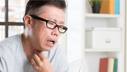 A person with glasses and a gray shirt touches their neck indoors, showing discomfort.
