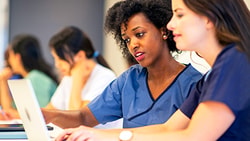 Two women in medical scrubs focus on a laptop screen, with others working in a medbridge classroom.