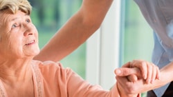 An older woman holds hands with a person in a blue shirt offering support, in a bright room.
