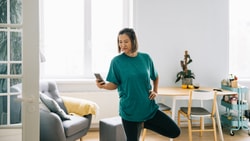 A woman in a teal shirt balances on one leg while looking at her phone in a living room near furniture.