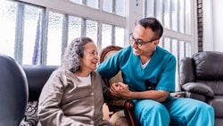 A male healthcare professional wearing teal scrubs and glasses sits next to an elderly woman, smiling warmly and holding her hand in a brightly lit room.